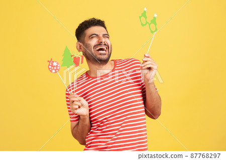 Overjoyed excited man with beard in striped t-shirt laughing out loud holding in hands holiday paper masks on stick, celebrating xmas and new year. Indoor studio shot isolated on yellow background 87768297
