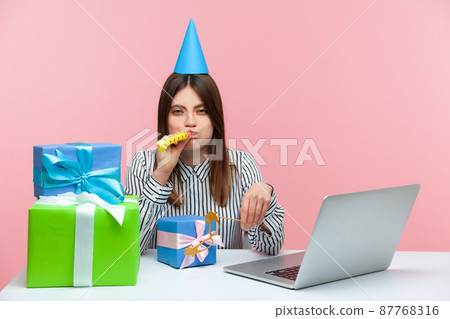 Unhappy brunette woman with party cone and blower sitting at workplace surrounded by many presents, feeling depressed celebrating birthday at work. Indoor studio shot isolated on pink background 87768316