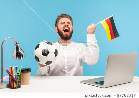Extremely excited bearded man football fan sincerely rejoicing, supporting favourite team holding ball and german flag, watching football at workplace. Indoor studio shot isolated on blue background 87768317