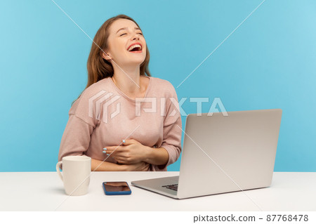Amused happy woman employee laughing out loud, holding her stomach and being hysterical at crazy joke on laptop, having break at workplace home office. indoor studio shot isolated on blue background 87768478