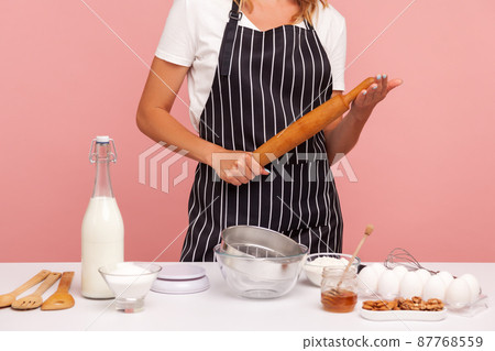 Cropped image of baker in striped apron, holding rolling pin in hands, being surrounded with ingredients for making pastry, cooking cakes. Indoor studio shot isolated on pink background. Cropped image of baker in striped apron, holding rolling pin in hands, being surrounded with ingredients for making pastry, cooking cakes. Indoor studio shot isolated on pink background. 87768559