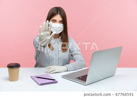 Quarantine bans. Woman office worker sitting safe healthy with hygienic face mask and protective gloves, showing stop gesture. Working in self-isolation, coronavirus outbreak. indoor, studio shot Quarantine bans. Woman office worker sitting safe healthy with hygienic face mask and protective gloves, showing stop gesture. Working in self-isolation, coronavirus outbreak. indoor, studio shot 87768591
