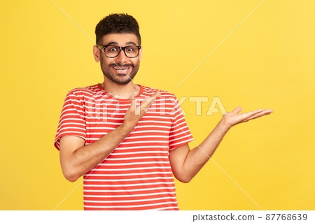 Advertise here. Happy excited bearded man in striped t-shirt showing empty space special for your advertisement, looking at camera with toothy smile. Indoor studio shot isolated on yellow background 87768639