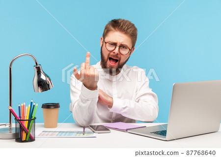 Excited rude man with beard office worker showing middle finger looking at camera sitting at workplace, uncultured gesture, disrespect and negativity. Indoor studio shot isolated on blue background 87768640
