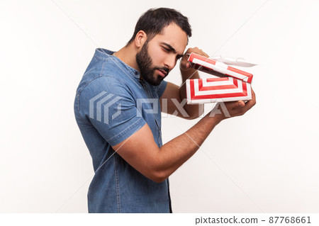 Side view nosy curious man with beard taking a look inside gift box, unpacking present to know what he received, frustrated with content. Indoor studio shot isolated on white background 87768661