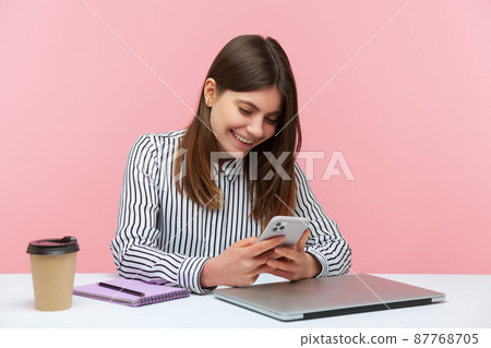 Smiling happy woman office worker in striped shirt chatting or surfing internet at smartphone sitting at workplace, pleasant conversation at messenger. Indoor studio shot isolated on pink background Smiling happy woman office worker in striped shirt chatting or surfing internet at smartphone sitting at workplace, pleasant conversation at messenger. Indoor studio shot isolated on pink background 87768705