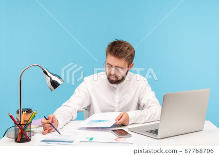 Concentrated business man in white shirt checking and analyzing information looking at graphs and diagrams, sitting at workplace with laptop. Indoor studio shot isolated on blue background Concentrated business man in white shirt checking and analyzing information looking at graphs and diagrams, sitting at workplace with laptop. Indoor studio shot isolated on blue background 87768706