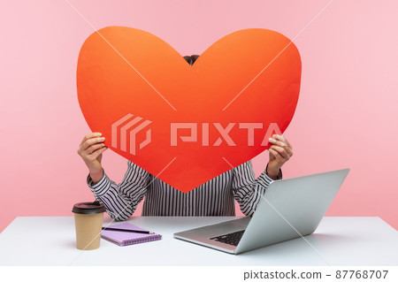 Woman hiding face behind huge red paper heart, using anonymous dating service on laptop, online chat and website. Indoor studio shot isolated on pink background Woman hiding face behind huge red paper heart, using anonymous dating service on laptop, online chat and website. Indoor studio shot isolated on pink background 87768707