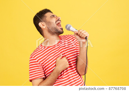 Side view positive bearded man in striped t-shirt loudly singing song holding microphone in hand, having fun resting in karaoke, singer performance. Indoor studio shot isolated on yellow background Side view positive bearded man in striped t-shirt loudly singing song holding microphone in hand, having fun resting in karaoke, singer performance. Indoor studio shot isolated on yellow background 87768708