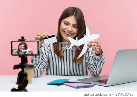 Smiling cheerful woman blogger showing credit card and paper plane on smartphone camera, recording video, giving tips about online booking. Indoor studio shot isolated on pink background Smiling cheerful woman blogger showing credit card and paper plane on smartphone camera, recording video, giving tips about online booking. Indoor studio shot isolated on pink background 87768755