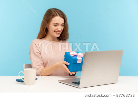 Beautiful woman siting at workplace alone and showing gift box to laptop screen, talking on video call, celebrating birthday alone on self quarantine. indoor studio shot isolated on blue background Beautiful woman siting at workplace alone and showing gift box to laptop screen, talking on video call, celebrating birthday alone on self quarantine. indoor studio shot isolated on blue background 87768758