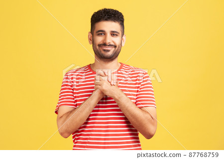 Happy positive man with beard in striped shirt pressing hands together on chest enjoying, calming and comforting himself. Indoor studio shot isolated on yellow background Happy positive man with beard in striped shirt pressing hands together on chest enjoying, calming and comforting himself. Indoor studio shot isolated on yellow background 87768759