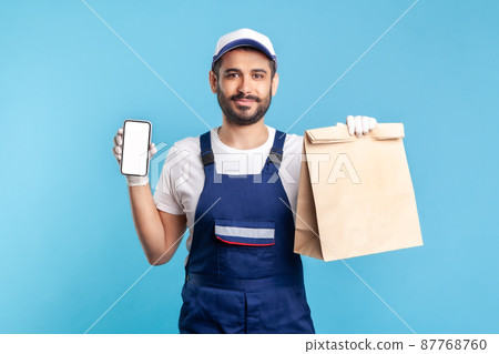 Delivery service mobile app. Courier man in uniform showing parcel and cellphone with blank display, mock up for express shipping advertise, online takeaway food order. indoor studio shot, isolated Delivery service mobile app. Courier man in uniform showing parcel and cellphone with blank display, mock up for express shipping advertise, online takeaway food order. indoor studio shot, isolated 87768760