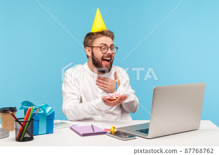Happy excited man office worker in party cone hat receiving congratulations online holding cake with candle before laptop camera sitting at workplace. Indoor studio shot isolated on blue background Happy excited man office worker in party cone hat receiving congratulations online holding cake with candle before laptop camera sitting at workplace. Indoor studio shot isolated on blue background 87768762