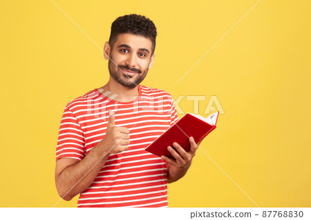Happy satisfied bearded man in red striped t-shirt showing thumbs up gesture holding and reading book, likes genre and plot. Indoor studio shot isolated on yellow background 87768830