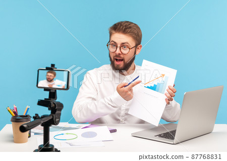 Male employee analyzing graph of clients growth, recording video on smartphone camera or streaming online, conducting conference, making report. Indoor studio shot isolated on blue background 87768831
