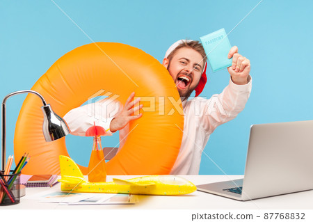Excited happy man office worker in santa claus hat sitting with toy plane, lemonade and in rubber ring, showing passport, going on winter vacation. Indoor studio shot isolated on blue background Excited happy man office worker in santa claus hat sitting with toy plane, lemonade and in rubber ring, showing passport, going on winter vacation. Indoor studio shot isolated on blue background 87768832