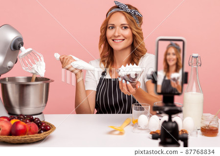 Blonde food blogger making video with tutorial of decoration desserts, looking at smart phone on tripod with smile, holding pastry cone with cream. Indoor studio shot isolated on pink background. Blonde food blogger making video with tutorial of decoration desserts, looking at smart phone on tripod with smile, holding pastry cone with cream. Indoor studio shot isolated on pink background. 87768834