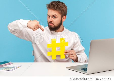 Serious bearded man smm manager pointing finger at hashtag sign, teaching social media management online, blog development. Indoor studio shot isolated on blue background Serious bearded man smm manager pointing finger at hashtag sign, teaching social media management online, blog development. Indoor studio shot isolated on blue background 87768835