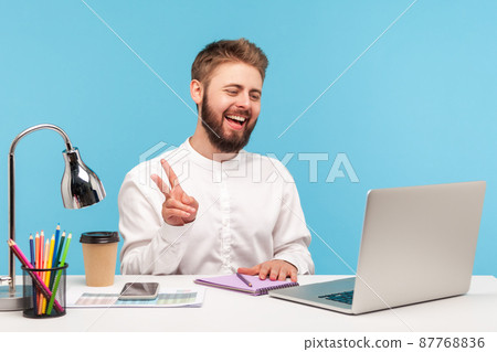 Positive bearded man office worker showing v gesture with fingers talking video call, looking at laptop camera with toothy smile sitting at workplace. Indoor studio shot isolated on blue background Positive bearded man office worker showing v gesture with fingers talking video call, looking at laptop camera with toothy smile sitting at workplace. Indoor studio shot isolated on blue background 87768836