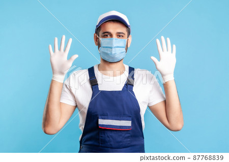 Cheerful handyman in overalls and mask raising hands with surgical gloves, smiling to camera. Safety clothes, protection and hygiene in service industry, courier delivery, housekeeping maintenance. Cheerful handyman in overalls and mask raising hands with surgical gloves, smiling to camera. Safety clothes, protection and hygiene in service industry, courier delivery, housekeeping maintenance. 87768839