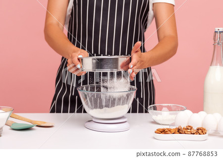 Cropped image of female confectioner sifts flour for doing homemade pastry, lady wearing apron, holds sieve in hands, surrounded with bakery ingredients. Indoor studio shot isolated on pink background Cropped image of female confectioner sifts flour for doing homemade pastry, lady wearing apron, holds sieve in hands, surrounded with bakery ingredients. Indoor studio shot isolated on pink background 87768853