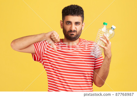 Sad unhappy bearded man showing thumbs down gesture holding in hand plastic bottles, dissatisfied with environmental pollution, worrying about ecology. Indoor studio shot isolated on yellow background 87768987