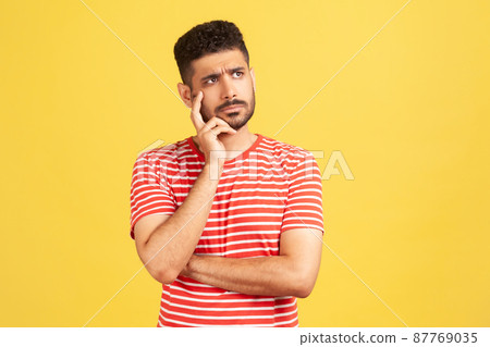 Doubtful bearded man in striped t-shirt contemplating leaning head on hand, pondering difficult question, trying to make right choice. Indoor studio shot isolated on yellow background Doubtful bearded man in striped t-shirt contemplating leaning head on hand, pondering difficult question, trying to make right choice. Indoor studio shot isolated on yellow background 87769035