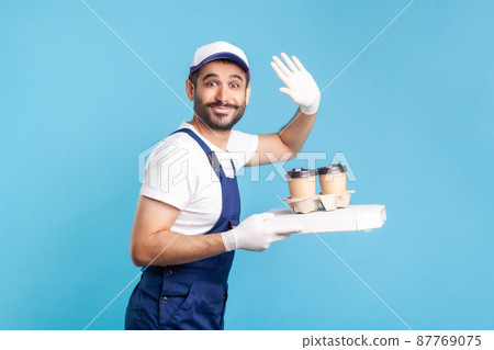 Delivery service. Amiable courier in overalls holding coffee and pizza box, wearing safety gloves offering drinks food and waving hello to camera. indoor studio shot isolated on blue background 87769075
