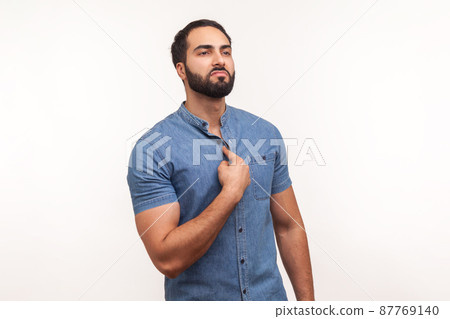 Self confident egoistic man with beard in blue shirt pointing finger at chest showing his strength and importance, proud of himself. Indoor studio shot isolated on white background Self confident egoistic man with beard in blue shirt pointing finger at chest showing his strength and importance, proud of himself. Indoor studio shot isolated on white background 87769140