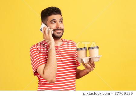 Busy bearded man in red striped t shirt taking phone holding in hand holder with two coffee cups, easy order fast delivery concept. Indoor studio shot isolated on yellow background Busy bearded man in red striped t shirt taking phone holding in hand holder with two coffee cups, easy order fast delivery concept. Indoor studio shot isolated on yellow background 87769159