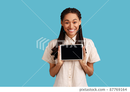 Smiling satisfied woman holds modern tablet with empty screen for your advertisement, poses with electronic gadget in hands, wearing white shirt. Indoor studio shot isolated on blue background. 87769164