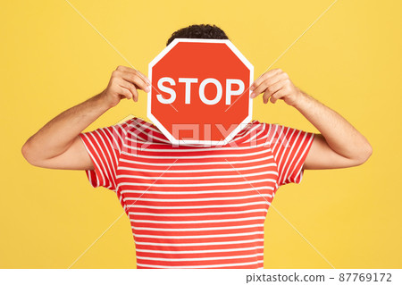 Unknown anonymous man in red striped t-shirt hiding his face behind red stop sign board, scared victim of humiliation and violence. Indoor studio shot isolated on yellow background Unknown anonymous man in red striped t-shirt hiding his face behind red stop sign board, scared victim of humiliation and violence. Indoor studio shot isolated on yellow background 87769172
