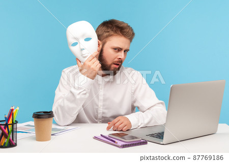 Unknown bearded man putting of white face mask sitting at workplace an looking at camera with mysterious facial expression, hiding his personality. Indoor studio shot isolated on blue background 87769186