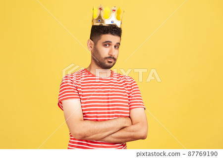 Arrogant selfish bearded man in striped t-shirt egoistically looking at camera, posing with crown on head, pretending to be king, privileged status. Indoor studio shot isolated on yellow background Arrogant selfish bearded man in striped t-shirt egoistically looking at camera, posing with crown on head, pretending to be king, privileged status. Indoor studio shot isolated on yellow background 87769190