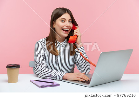 Excited positive secretary in striped shirt making calls on landline telephone working on laptop sitting at office, looking at camera with toothy smile. Indoor studio shot isolated on pink background Excited positive secretary in striped shirt making calls on landline telephone working on laptop sitting at office, looking at camera with toothy smile. Indoor studio shot isolated on pink background 87769216