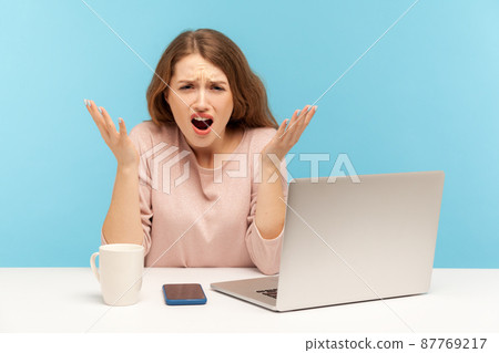 How could you. Annoyed young woman employee raising hands in indignant gesture, asking why and looking angrily, sitting at workplace with laptop. indoor studio shot isolated on blue background How could you. Annoyed young woman employee raising hands in indignant gesture, asking why and looking angrily, sitting at workplace with laptop. indoor studio shot isolated on blue background 87769217