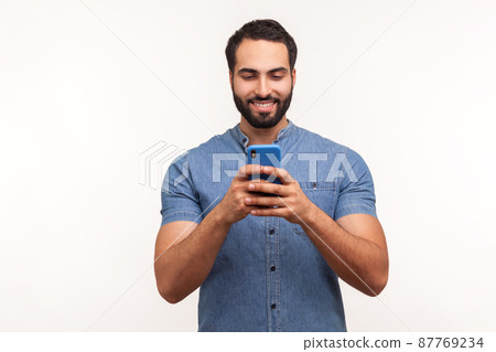 Smiling positive bearded man in blue shirt looking at smartphone display, surfing internet, betting, doing shopping online. Indoor studio shot isolated on white background 87769234