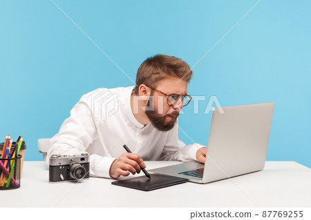 Concentrated bearded man designer drawing on professional tablet looking at laptop display, creating project sitting at workplace. Indoor studio shot isolated on blue background 87769255