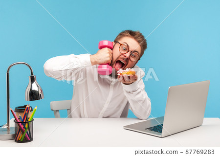 Funny crazy man office worker holding dumbbell but wanting to eat sweet donut ignoring training, temptation, unhealthy eating during work. Indoor studio shot isolated on blue background 87769283