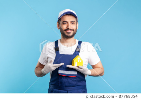 Portrait of happy handyman, mover guy in workwear and gloves showing paper house. Advertising home repair, maintenance or moving services, help in relocation. studio shot isolated on blue background Portrait of happy handyman, mover guy in workwear and gloves showing paper house. Advertising home repair, maintenance or moving services, help in relocation. studio shot isolated on blue background 87769354