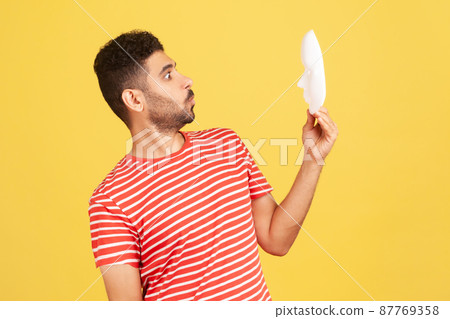 Profile portrait of bearded man in striped t-shirt looking at white mask with unknown face with surprised expression, trying to guess emotion. Indoor studio shot isolated on yellow background Profile portrait of bearded man in striped t-shirt looking at white mask with unknown face with surprised expression, trying to guess emotion. Indoor studio shot isolated on yellow background 87769358