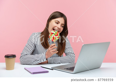 Funny playful woman licking and biting round colored lollipop on stick talking on video call at laptop sitting at workplace, having fun. Indoor studio shot isolated on pink background 87769359
