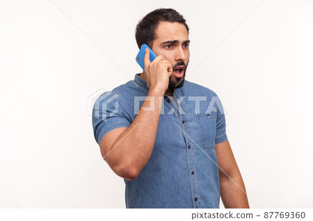 Shocked man with beard in blue shirt talking phone, surprised with news looking big eyes and widely opened mouth, unpleasant call, bad tariffs, roaming. Indoor studio shot isolated on white background Shocked man with beard in blue shirt talking phone, surprised with news looking big eyes and widely opened mouth, unpleasant call, bad tariffs, roaming. Indoor studio shot isolated on white background 87769360