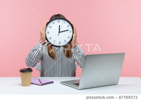 Time to work. Woman office worker sitting at workplace hiding face behind big wall clock, time management, schedule and business meeting appointment. Indoor studio shot isolated on pink background Time to work. Woman office worker sitting at workplace hiding face behind big wall clock, time management, schedule and business meeting appointment. Indoor studio shot isolated on pink background 87769372