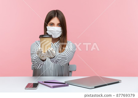 Woman office employee showing cup of coffee, sitting at workplace, feeling safe healthy with hygienic face mask and protective gloves, working in self-isolation, coronavirus quarantine. studio shot 87769379