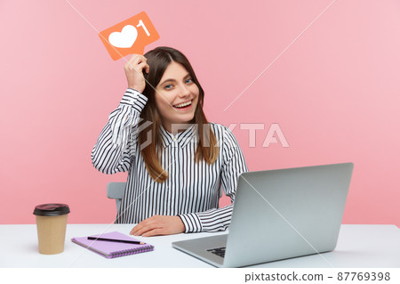 Excited female blogger holding likes counter template above head sitting at workplace with laptop, making posts in social networks. Indoor studio shot isolated on pink background 87769398