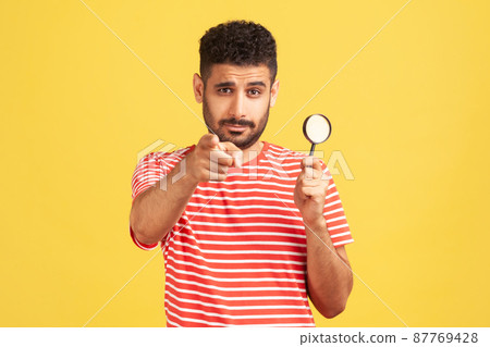 Serious confident man private detective in striped t-shirt holding in hand magnifying glass and pointing finger on you looking at camera, spying. Indoor studio shot isolated on yellow background 87769428