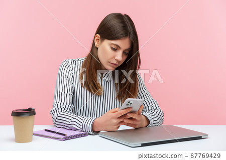 Serious concentrated business woman in striped shirt keeping business correspondence on her smartphone sitting at workplace, blogging. Indoor studio shot isolated on pink background Serious concentrated business woman in striped shirt keeping business correspondence on her smartphone sitting at workplace, blogging. Indoor studio shot isolated on pink background 87769429