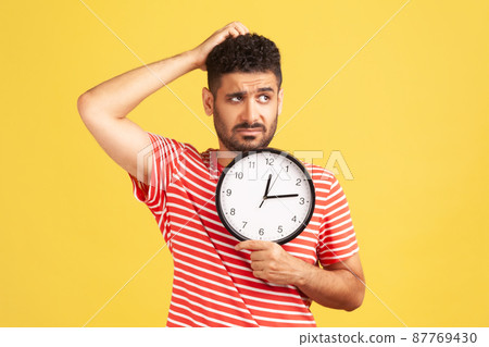 Confused thoughtful man with beard in red striped t-shirt rubbing his head holding in hands big wallclock, time out, worried about deadline. Indoor studio shot isolated on yellow background Confused thoughtful man with beard in red striped t-shirt rubbing his head holding in hands big wallclock, time out, worried about deadline. Indoor studio shot isolated on yellow background 87769430
